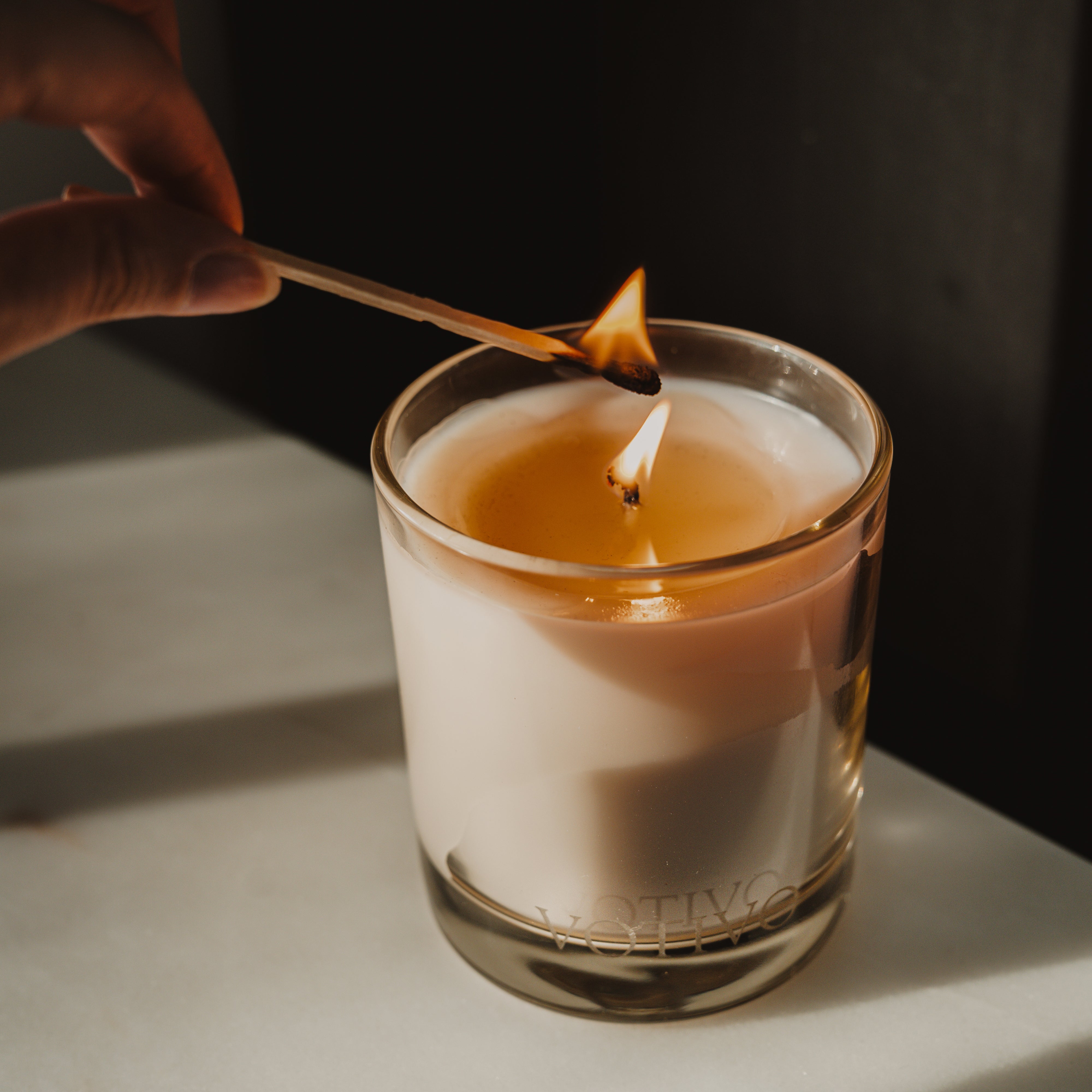 A hand lights the Votivo Red Currant 10 oz. Signature Candle in a glass container. Its soy blend wax burns brightly, illuminating the VOTIVO label and filling the space with a warm, inviting glow.