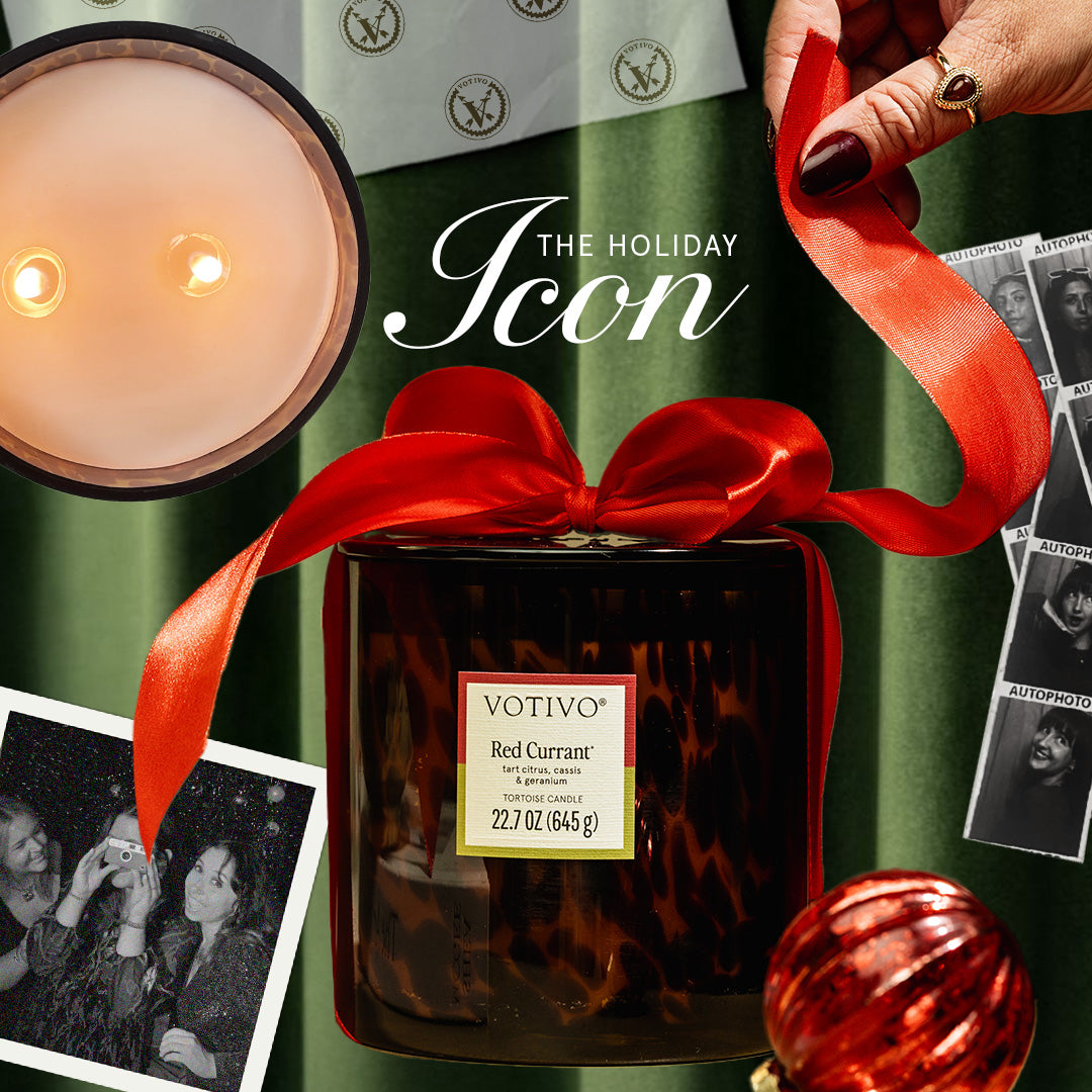 A hand ties a red ribbon around a Votivo Red Currant Tortoise Candle. The candle sits on a green surface along with a red ornament, black-and-white photos, and a lit candle. Text reads The Holiday Icon.
