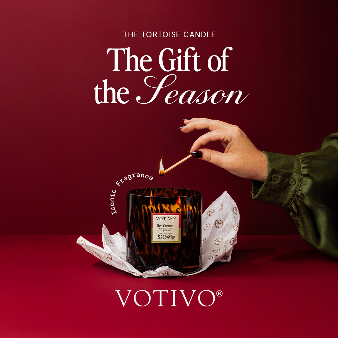 A hand lights the Votivo Red Currant Tortoise Candle, a soy blend in a tortoise-patterned glass jar. Set against a deep red backdrop with "The Gift of the Season" above, wrapping paper partially reveals its signature red currant scent.