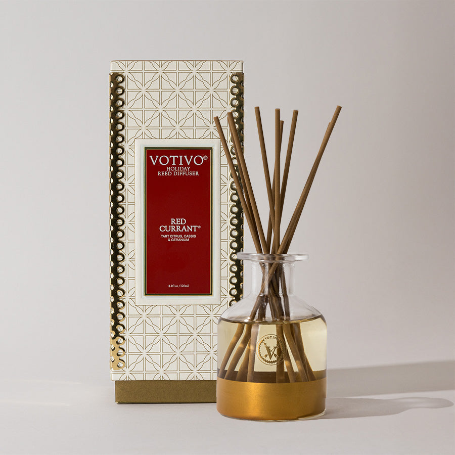 The Votivo Red Currant Holiday Reed Diffuser features a glass bottle with yellow liquid, several reeds, and comes in a festive box with gold and red accents, all in Votivos signature Red Currant fragrance.