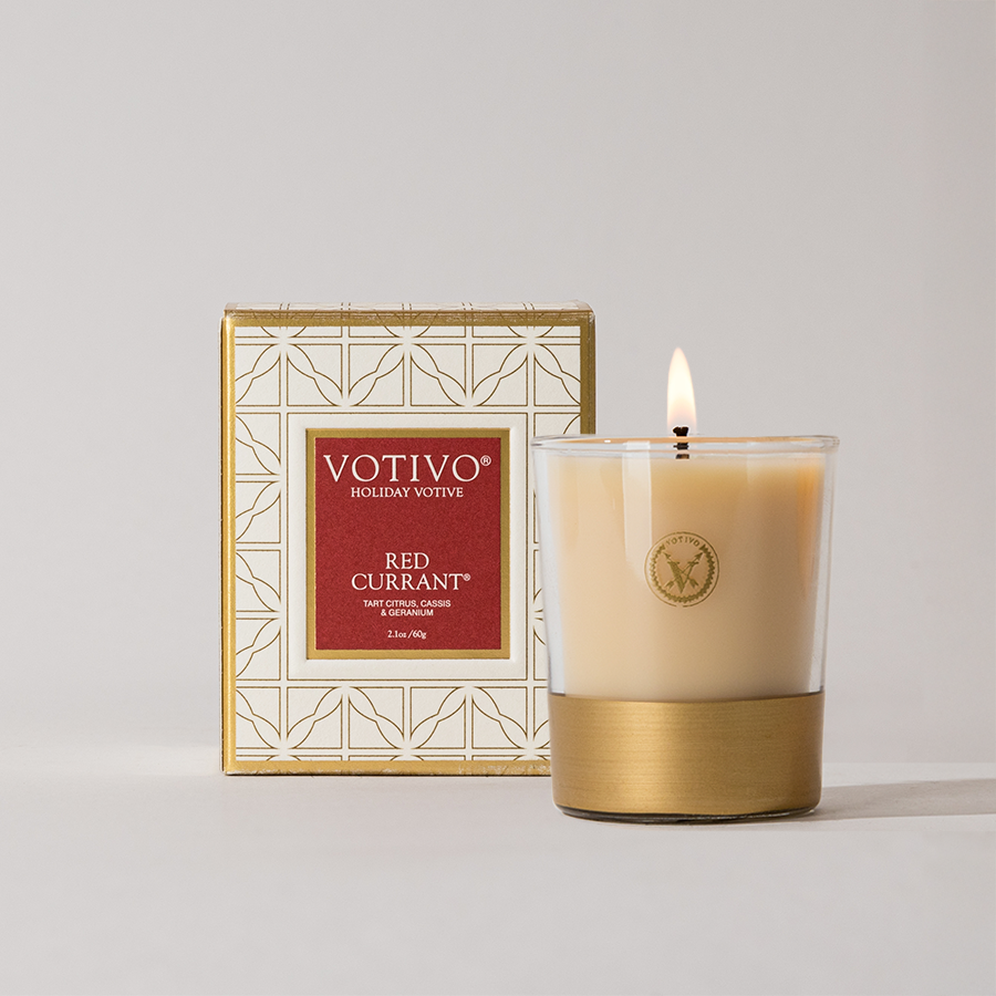 The Votivo Red Currant Holiday Votive candle glows in a glass holder with a gold base, beside its geometric gold-and-white box—a luxurious gift featuring a vibrant fragrance and elegant holiday style.