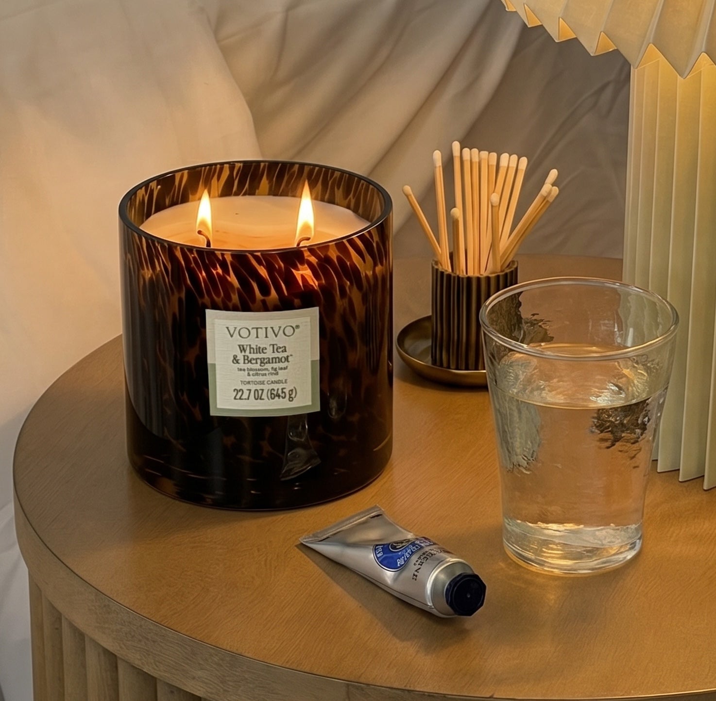 A lit Votivo White Tea & Bergamot Tortoise Candle crafted from soy blend wax, a tube of hand cream, a glass of water, and a cup of cotton swabs sit on a wooden nightstand next to a lamp with a pleated shade and draped fabric in the background. The gentle aroma of white tea and bergamot lingers softly in the air.