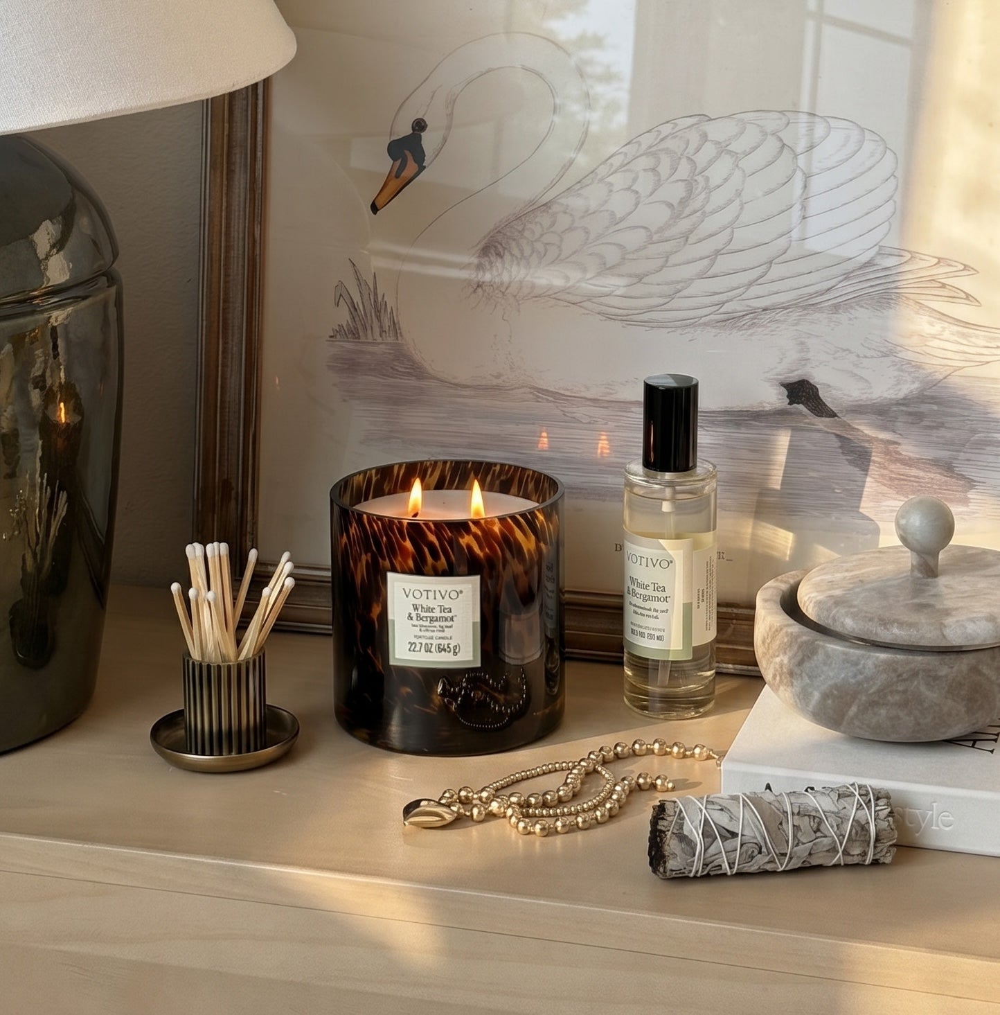A cozy tabletop scene featuring the Votivo White Tea & Bergamot Tortoise Candle lit beside a spray bottle, matches, beaded bracelet, sage, marble container, and framed swan art. Warm sunlight streams in.
