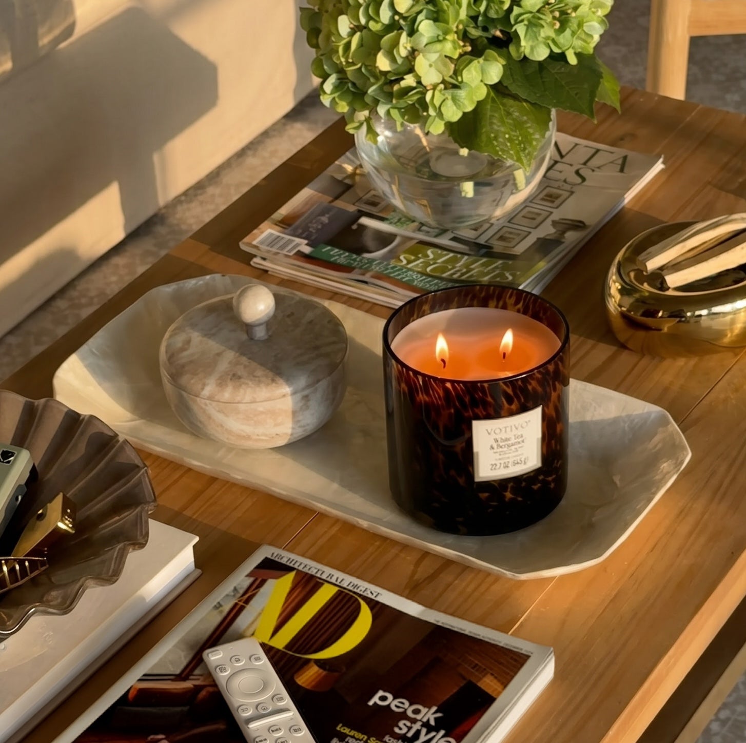 A wooden coffee table displays the Votivo White Tea & Bergamot Tortoise Candle, crafted from a soy blend wax and lit beside a marble jar, magazines, a remote, a decorative leaf tray, and a vase of green hydrangeas—all arranged neatly in warm sunlight.