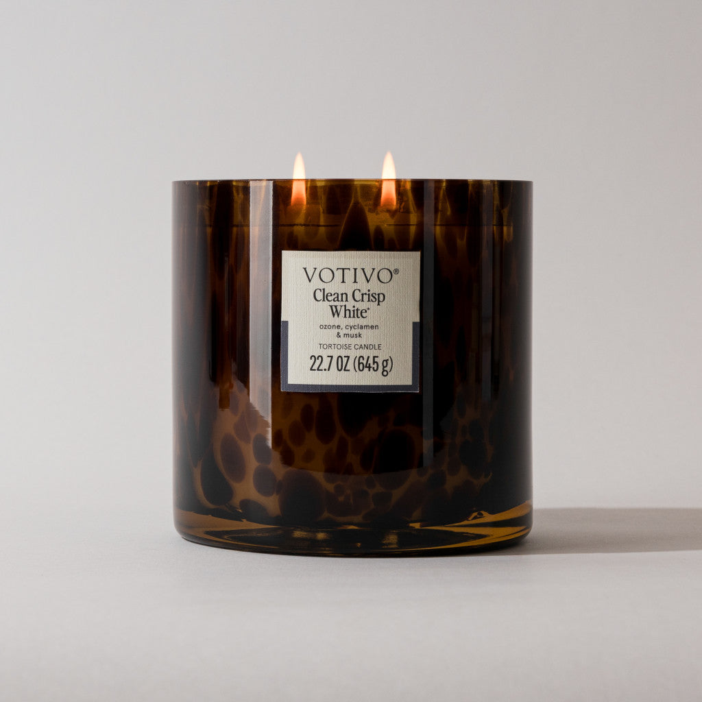 A lit Votivo Clean Crisp White Tortoise Candle sits on a plain white surface, its brown speckled jar featuring the brand, product name, fresh scent details, and candle weight (22.7 oz / 645 g).