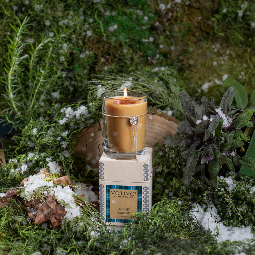 The Votivo Wilde 6.8 oz. Aromatic Candle, in its glass holder and box, is set among green moss, herbs, and snowy patches for a rustic look that highlights its unique fragrance.