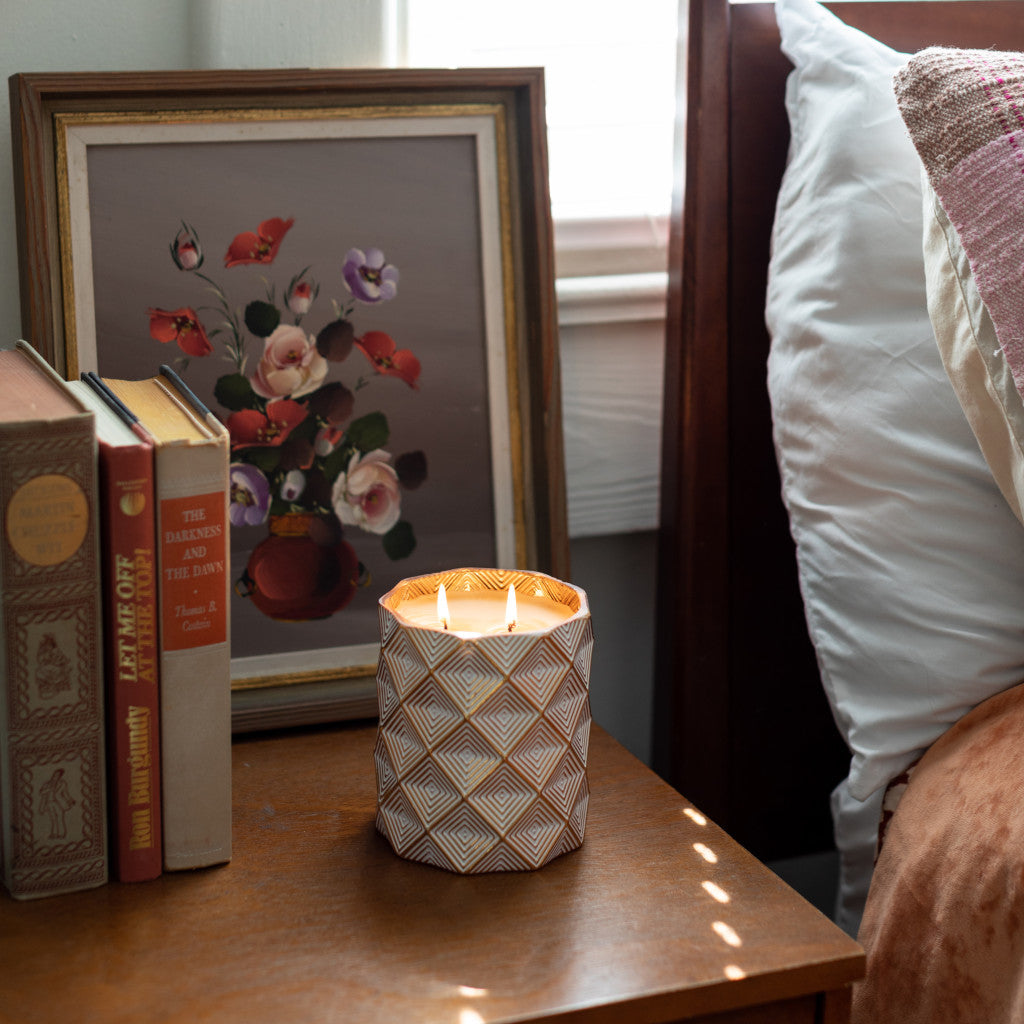 The Votivo Icy Blue Pine Holiday Decorative Candle in a patterned holder sits on a bedside table beside books and a floral painting, with part of a bed visible as sunlight highlights its festive scent.