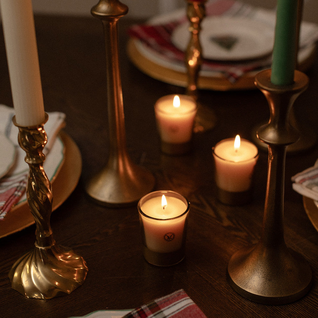 A cozy table features Votivo’s Spiced Chai Holiday Votive, brass holders, plaid napkins, and plates with gold chargers—creating a warm atmosphere as the soy blend wax fills the room with an inviting festive fragrance.