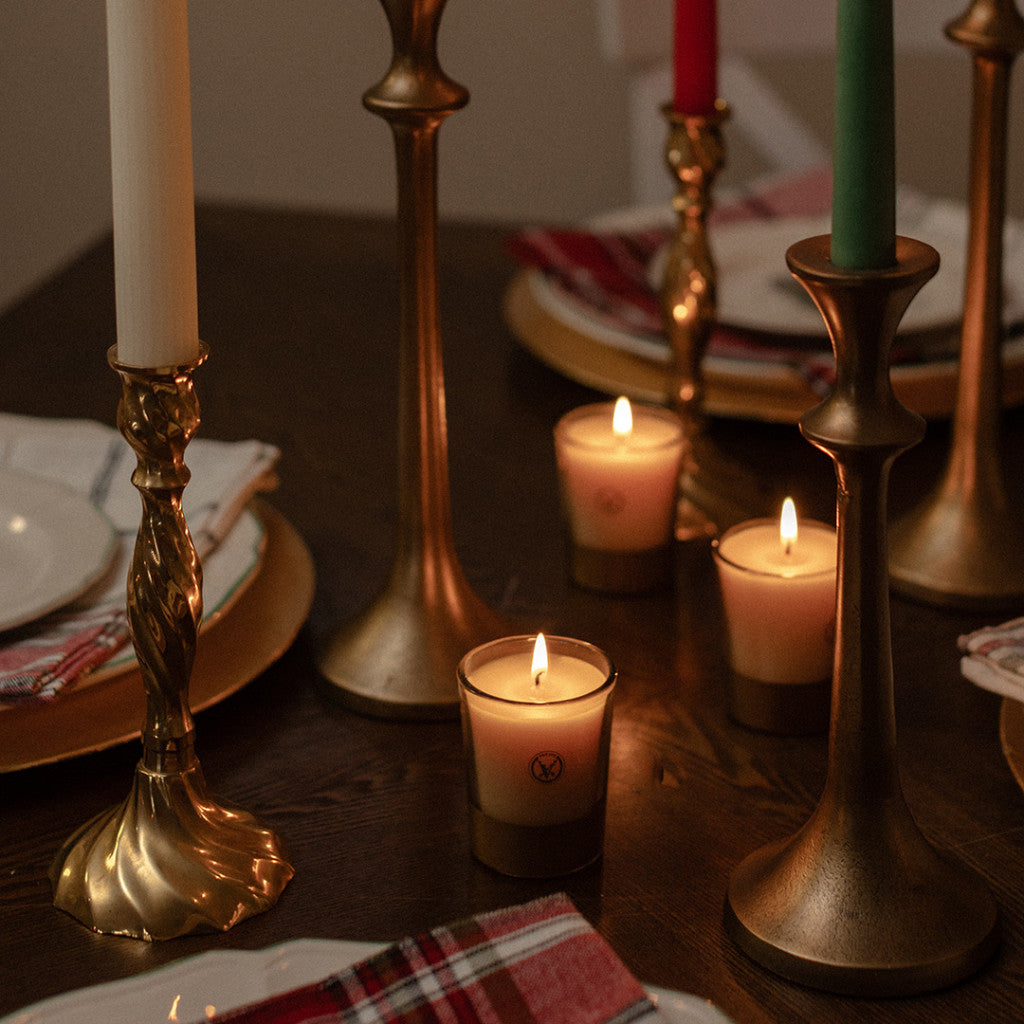 A close-up of a dining table set with gold candle holders, lit white candles, plaid napkins, and white plates, featuring the Votivo Icy Blue Pine Holiday Votive made from soy blend wax for a warm, festive atmosphere.