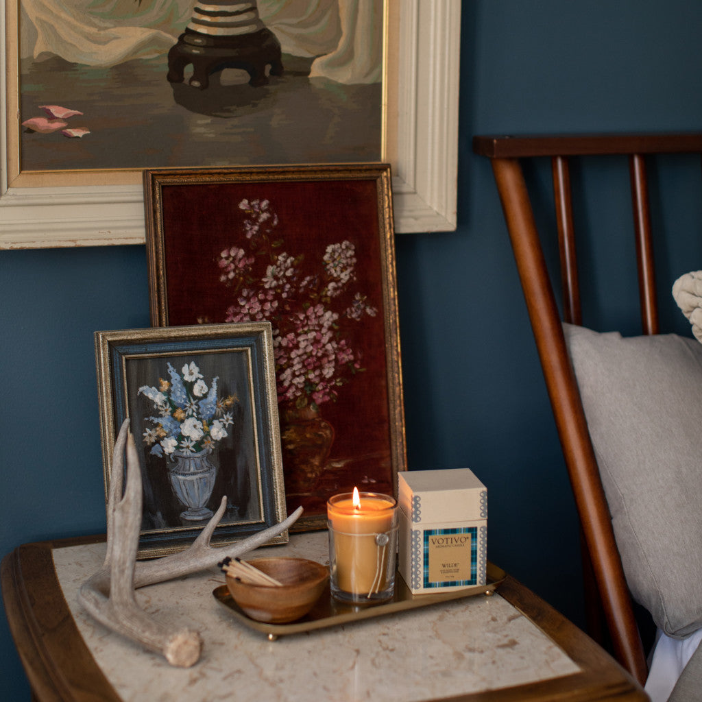 A wooden nightstand with a tray holds the Votivo Wilde 6.8 oz. Aromatic Candle, a lidded wooden bowl, and antler decor. Two framed floral paintings lean against a blue wall beside a wooden chair with a gray cushion.
