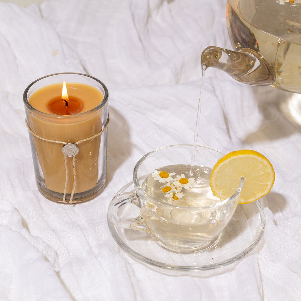 Hot water pours from a teapot into a glass cup with chamomile flowers and lemon. Next to it, the Votivo White Tea & Bergamot 6.8 oz aromatic candle in a glass holder rests on white, wrinkled cloth.