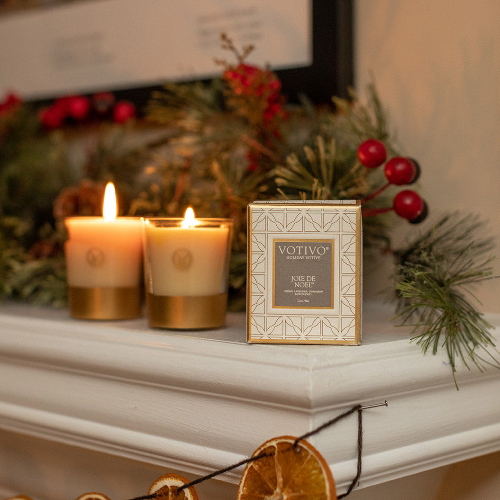 Two lit luxury candles and a Votivo Joie de Noel Holiday Votive sit on a pine-and-berry adorned mantel with dried orange garland. The soy blend wax of the Votivo candle fills the room with a warm, festive scent.