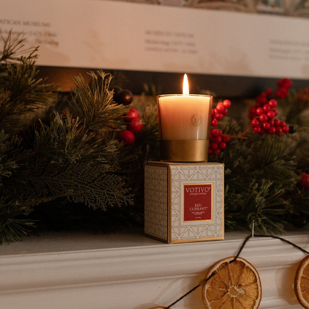 The Votivo Red Currant Holiday Votive sits on a white surface, surrounded by festive greenery, red berries, and dried orange slices for a cozy holiday ambiance.