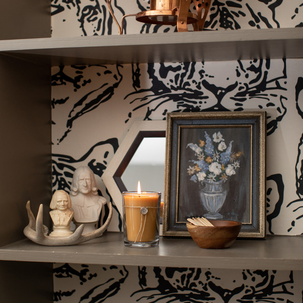 A shelf with the Votivo Wilde 6.8 oz. Aromatic Candle, a bowl of matches, a framed flower painting, a small mirror, antlers, and two sculpture busts are arranged against black and beige patterned wallpaper.