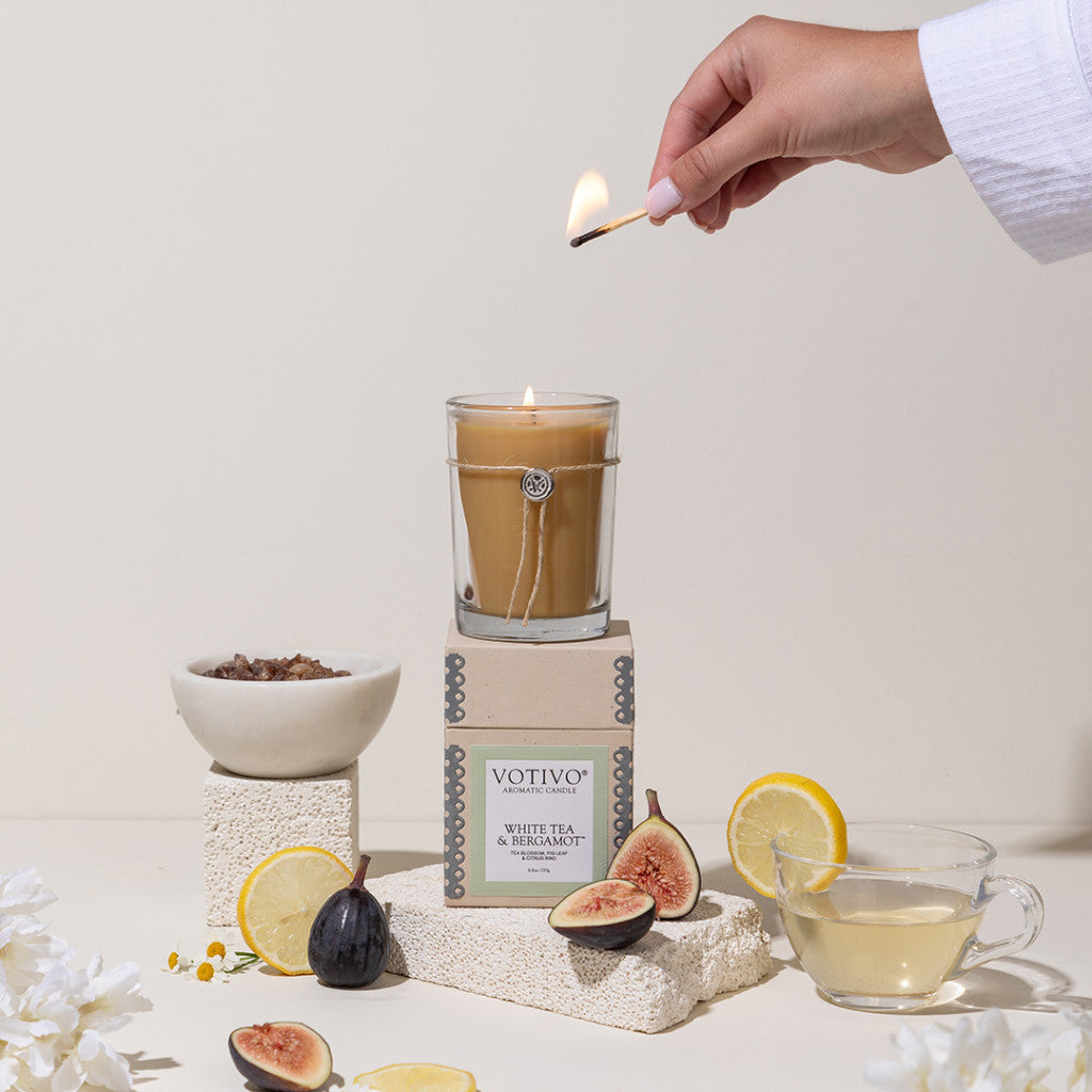 A hand lights the Votivo White Tea & Bergamot 6.8 oz Aromatic Candle, crafted from a soy blend, displayed on stacked boxes with figs, lemons, loose tea, a teacup, and white flowers arranged on a light background.