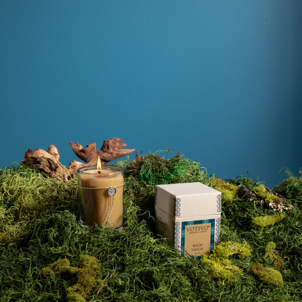 The Votivo Wilde 6.8 oz. Aromatic Candle, crafted from a premium soy blend for a clean, unique scent, glows in a glass holder atop green moss beside its beige box, with driftwood and a blue wall in the background.