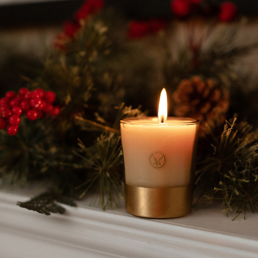The Votivo Red Currant Holiday Votive sits in a glass holder with a gold base on a white surface, surrounded by pine branches, red berries, and a pinecone for a festive holiday atmosphere.