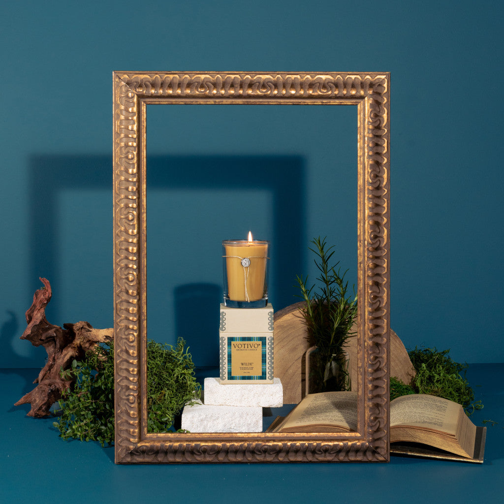 The Votivo Wilde 6.8 oz. Aromatic Candle, crafted from a soy blend, rests on stacked white blocks with greenery and open books nearby, all within an ornate gold frame set against a blue background.