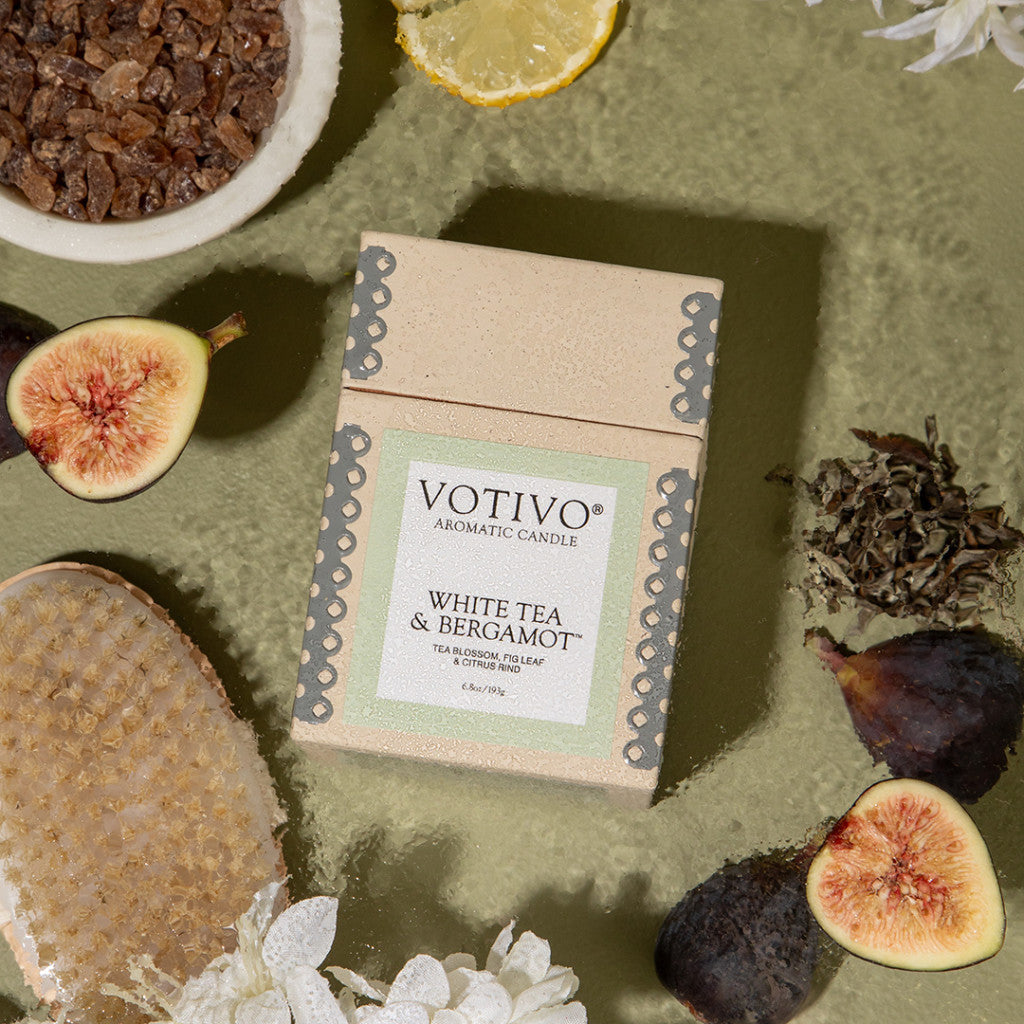 A Votivo White Tea & Bergamot 6.8 oz Aromatic Candle sits on a green surface, with figs, a lemon slice, dried tea leaves, brown sugar, a body brush, and white flowers. Made with soy blend wax for a clean, luxurious burn.