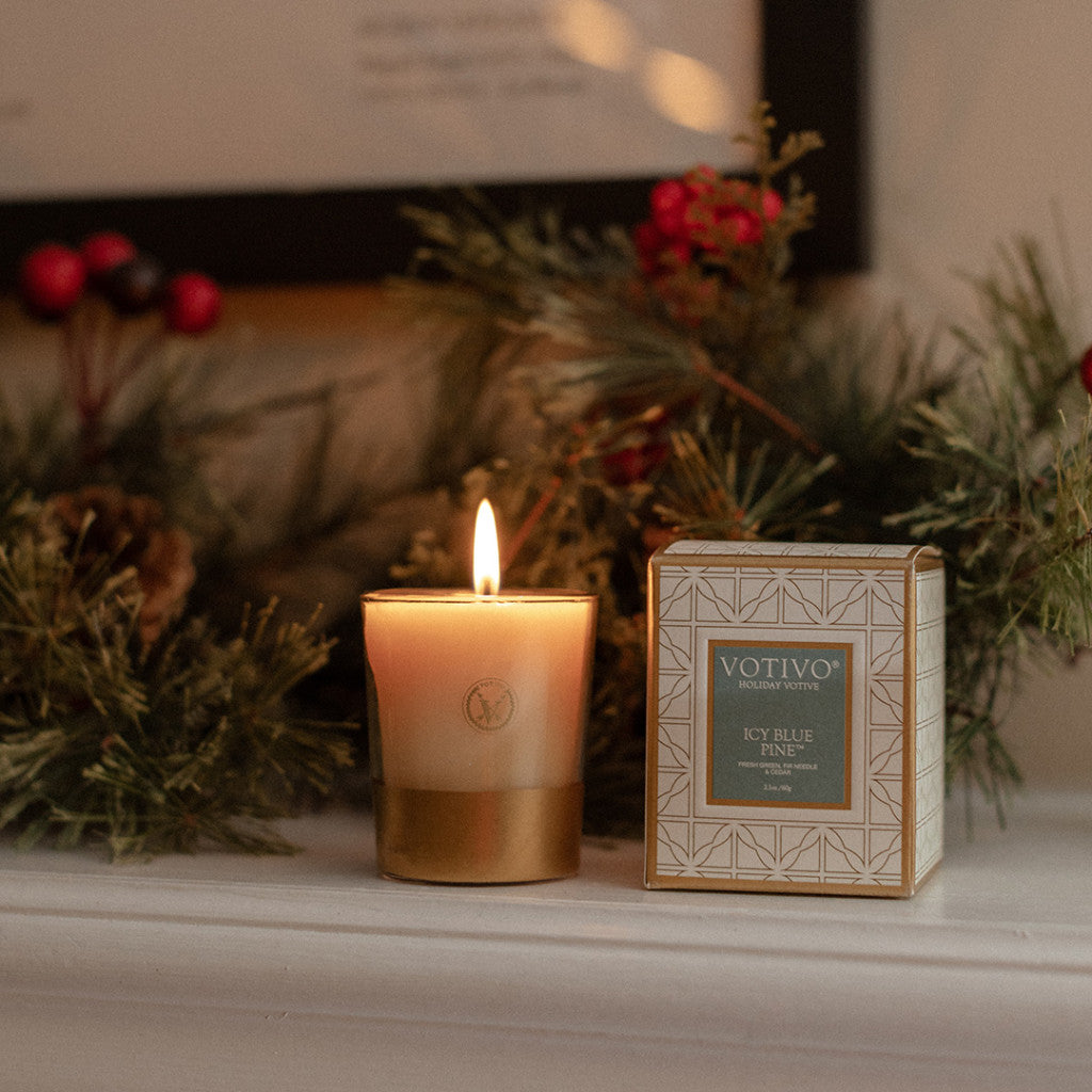 A glowing Votivo Icy Blue Pine Holiday Votive in a gold holder rests on a pine and berry–trimmed mantel, filling the room with its crisp soy blend scent.