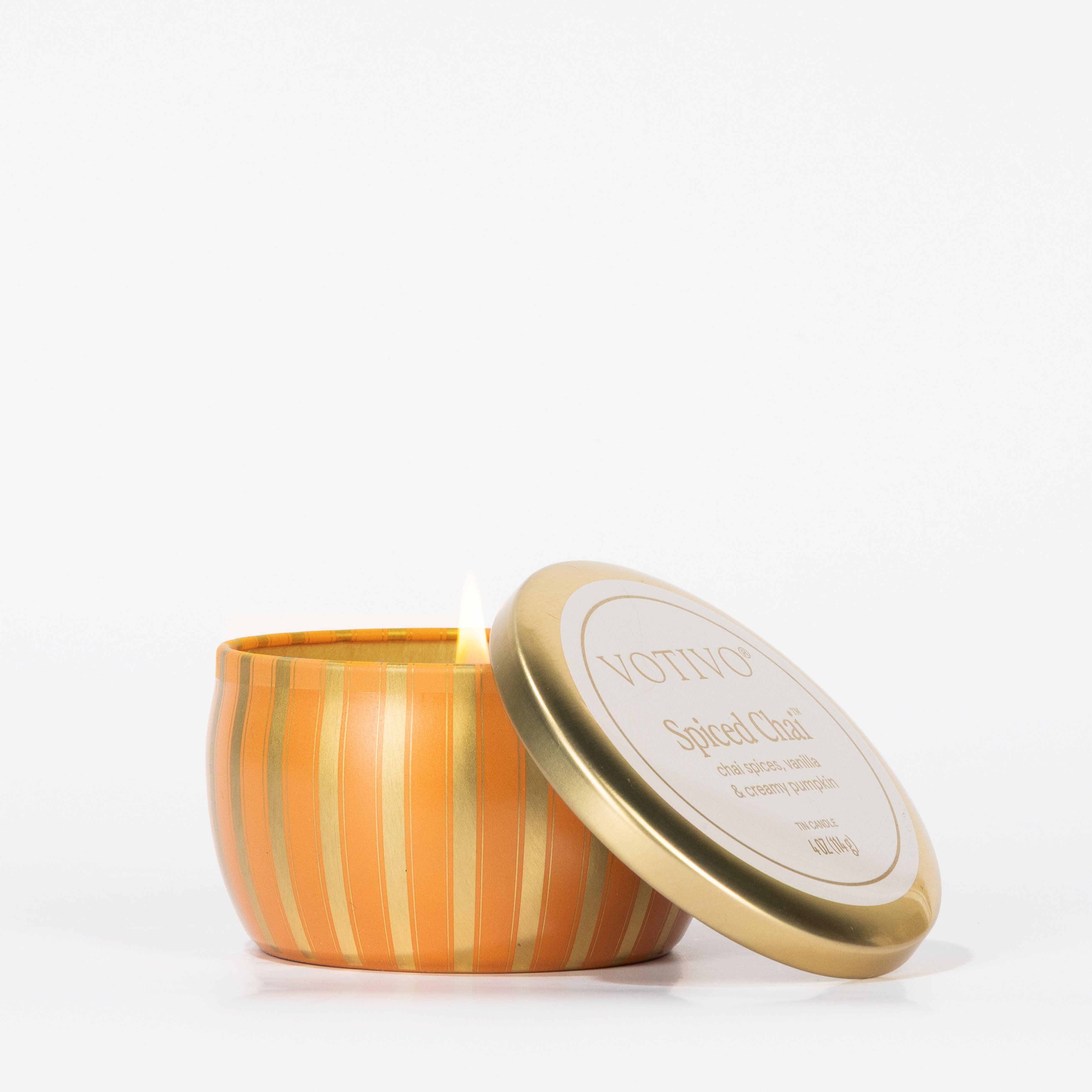 A Votivo Spiced Chai Holiday Tin Candle with orange and gold stripes sits on a white background, its lid partially open to reveal a lit soy blend wax candle inside.