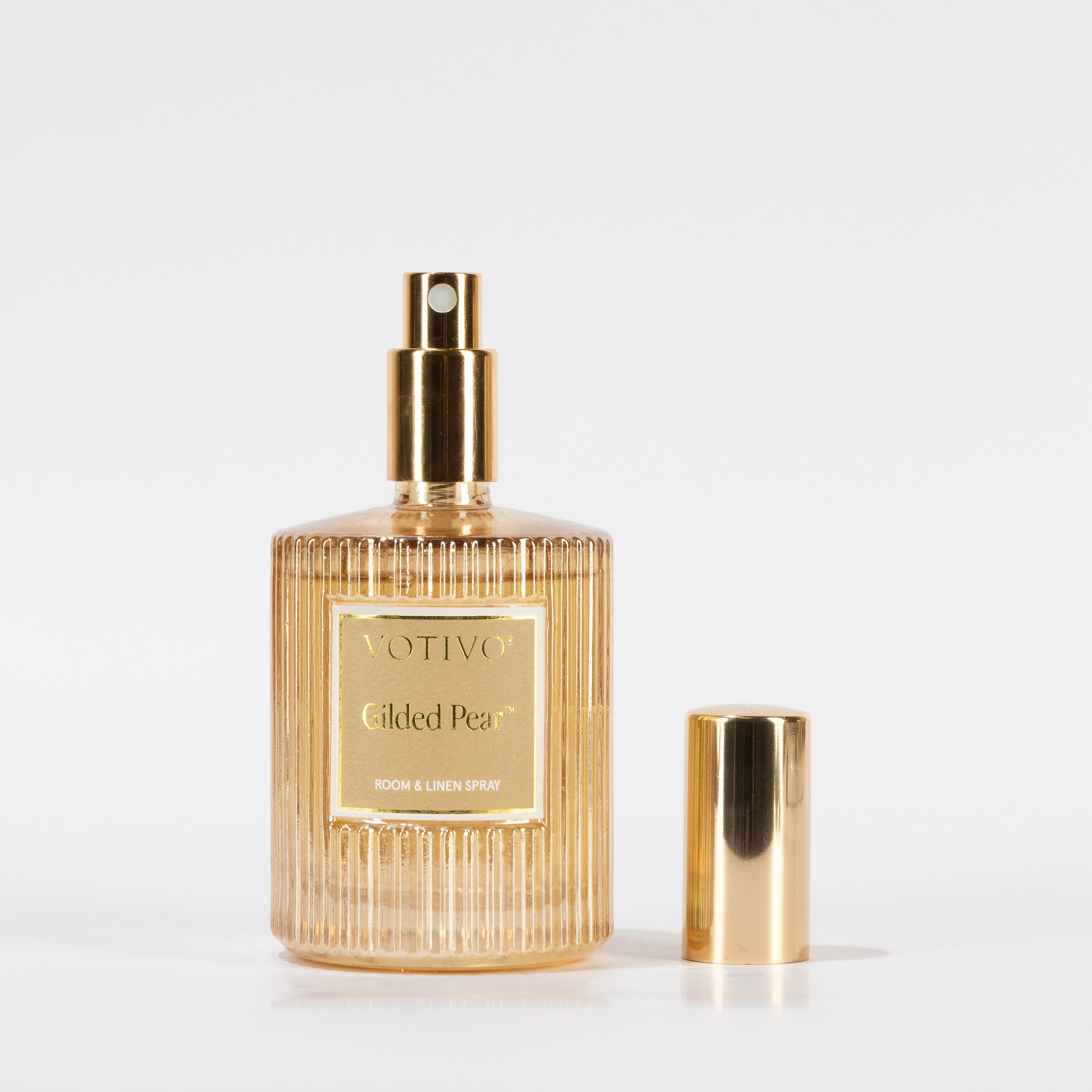 A clear, ribbed glass bottle of Votivo Gilded Pear Room & Linen Spray with a gold label and spray nozzle stands on a white background, its matching gold cap placed to the right. Safe for use on fabric.