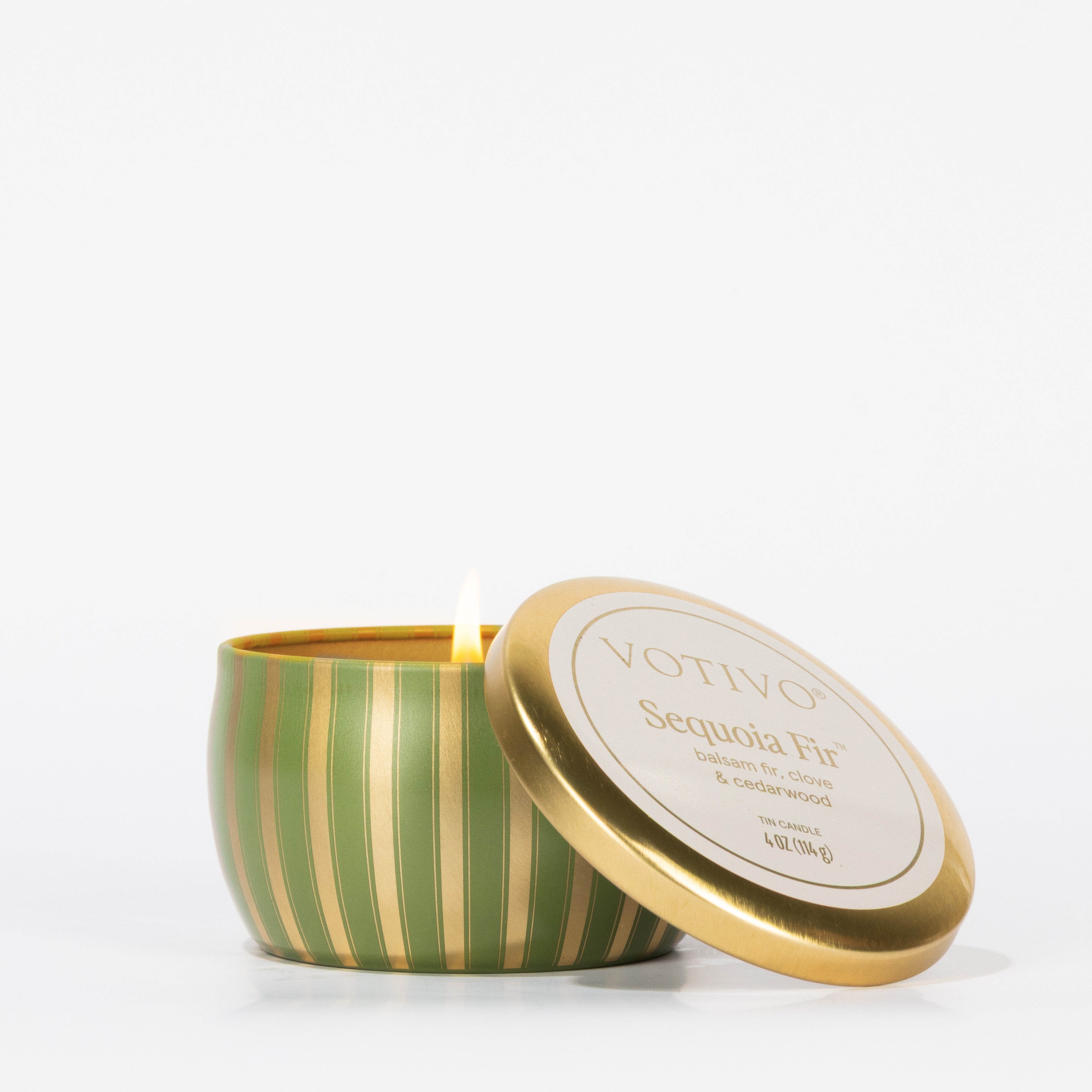 A lit Votivo Sequoia Fir Holiday Tin Candle in a green and gold striped tin, lid set aside, sits against a plain white background. The label displays the brand and scent name.