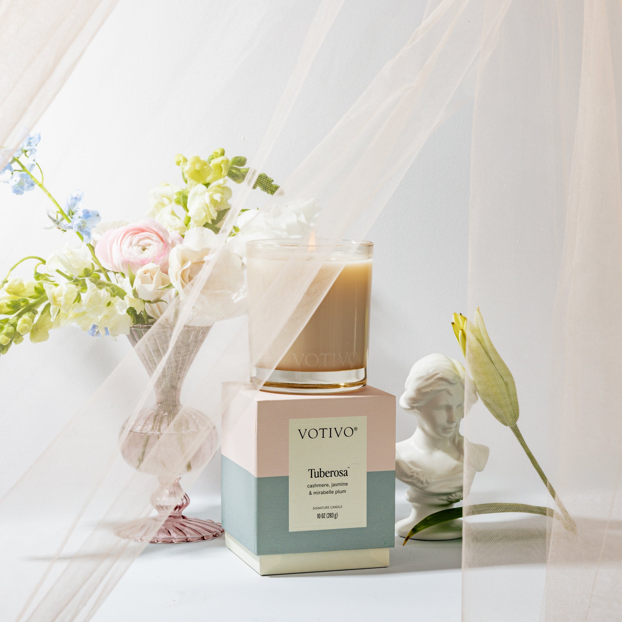 A Votivo Tuberosa 10 oz. Signature Candle made from soy blend wax rests on its box next to pastel flowers and a white bust, all softly draped in sheer fabric and gentle natural light.