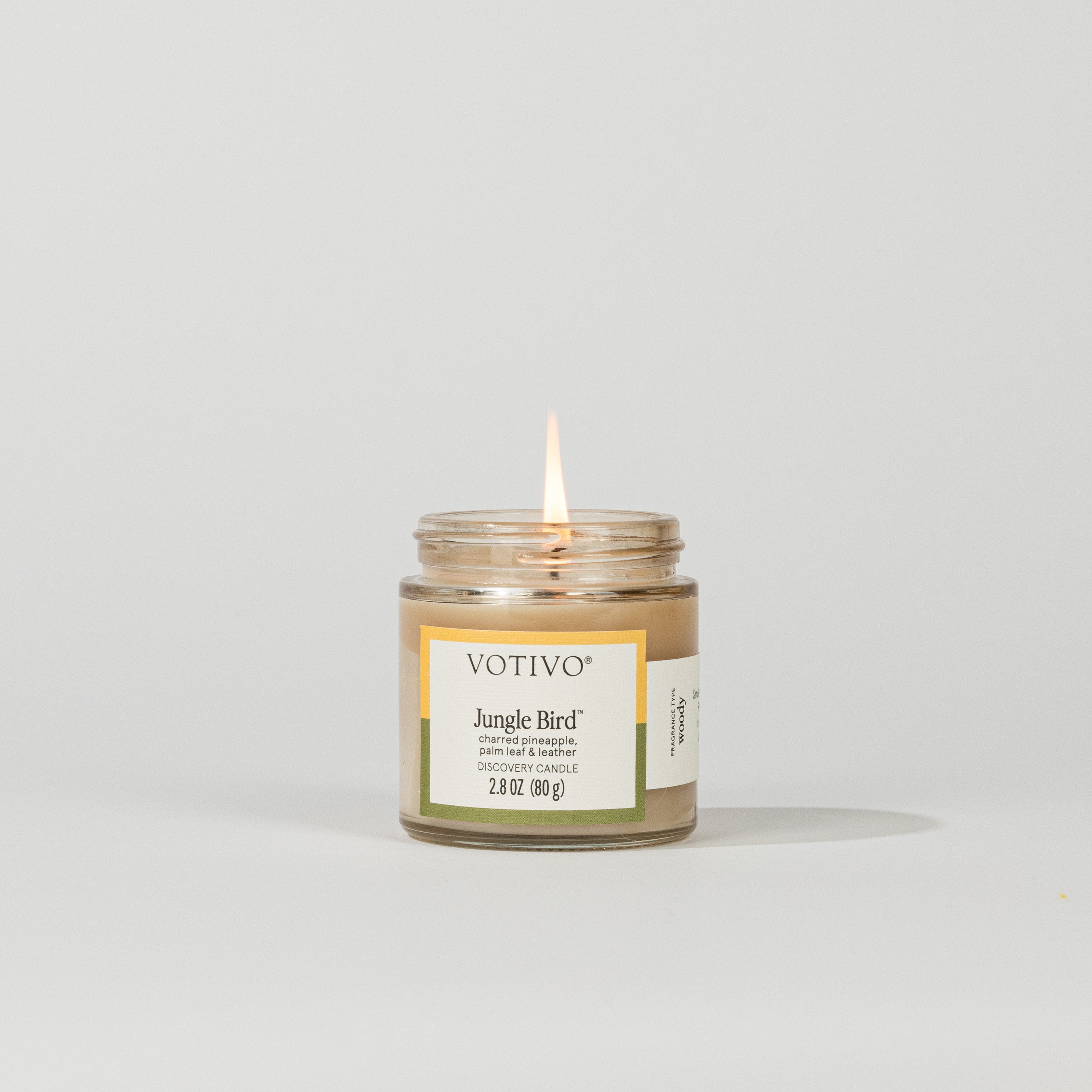 A small Votivo Jungle Bird Discovery Candle burns with a single flame against a white background. This soy blend candle has a white and green label and contains 2.8 oz (80g) of long-lasting fragrance.