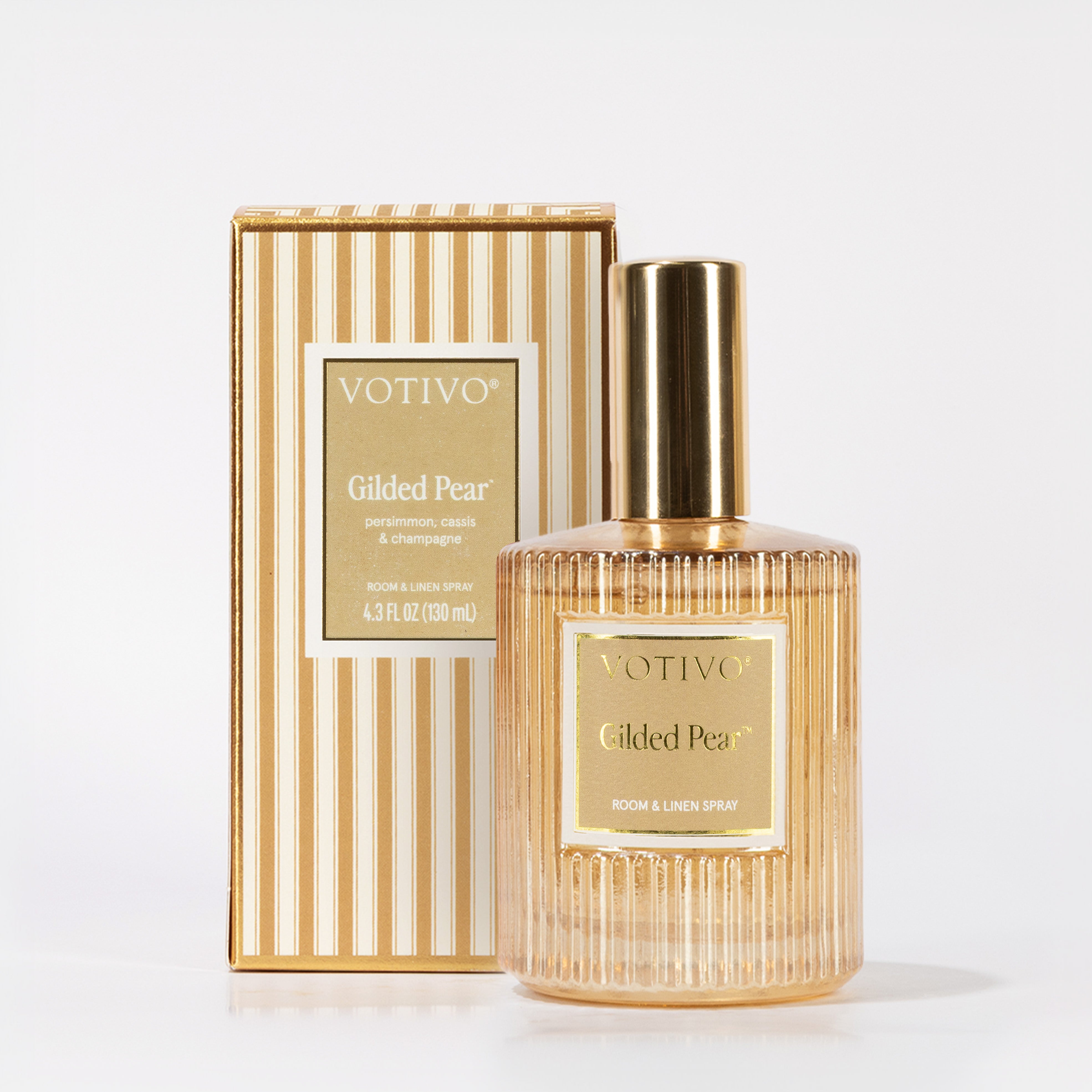 The Votivo Gilded Pear Room & Linen Spray, safe for fabrics, stands before its striped box—both labeled with scent notes of persimmon, cassis, and champagne. The elegant spray comes in a gold ribbed bottle design.