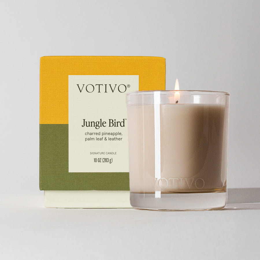 The Votivo Jungle Bird 10 oz. Signature Candle, made from soy blend wax, glows in a clear glass jar beside its box and delivers a unique scent of charred pineapple, palm leaf, and leather.