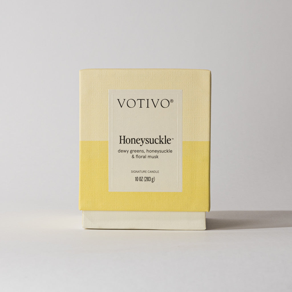 The Votivo Honeysuckle 10 oz. Signature Candle features soy blend wax, dewy greens, honeysuckle, and floral musk notes. It comes in a cream and yellow box with black text on a plain light background.