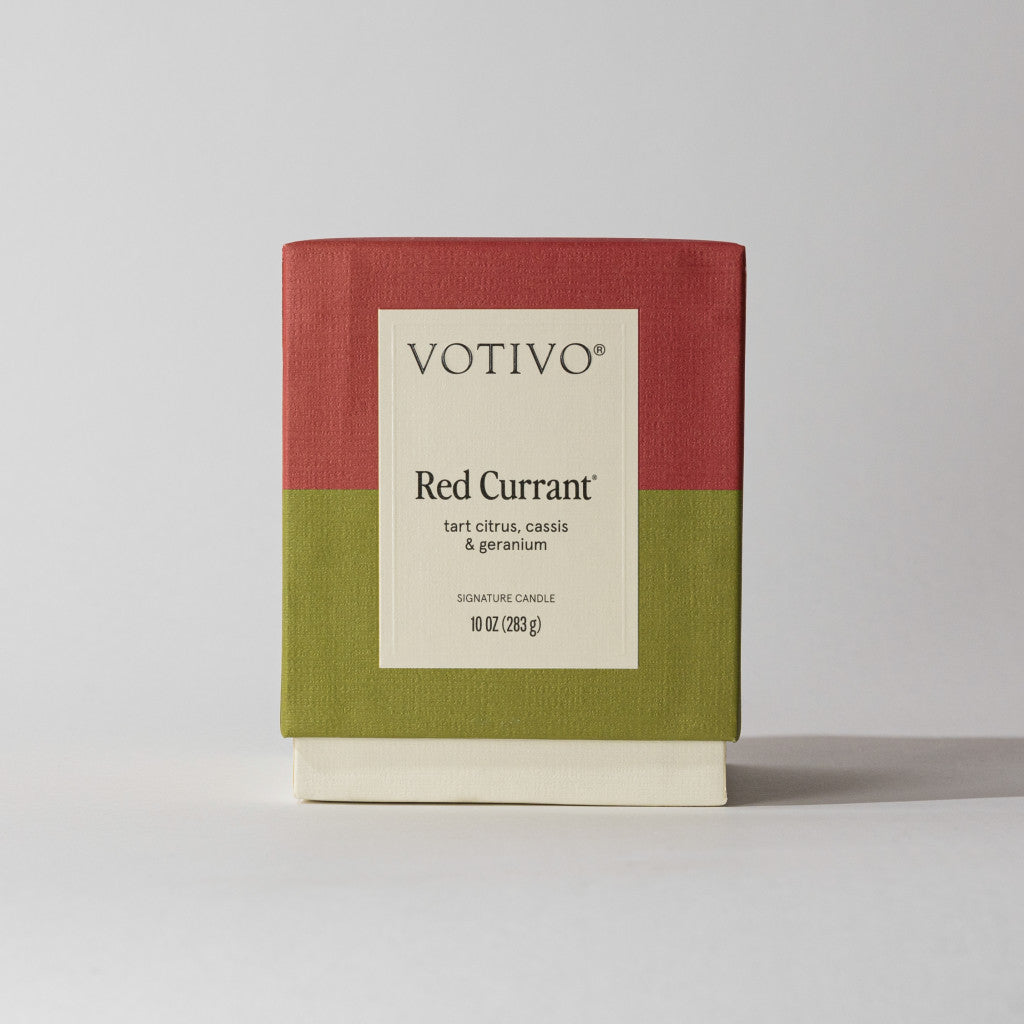 The Votivo Red Currant 10 oz. Signature Candle features a red top, olive green base, and cream label displaying tart citrus, cassis, and geranium scent notes. Made with premium soy blend wax for a clean burn.