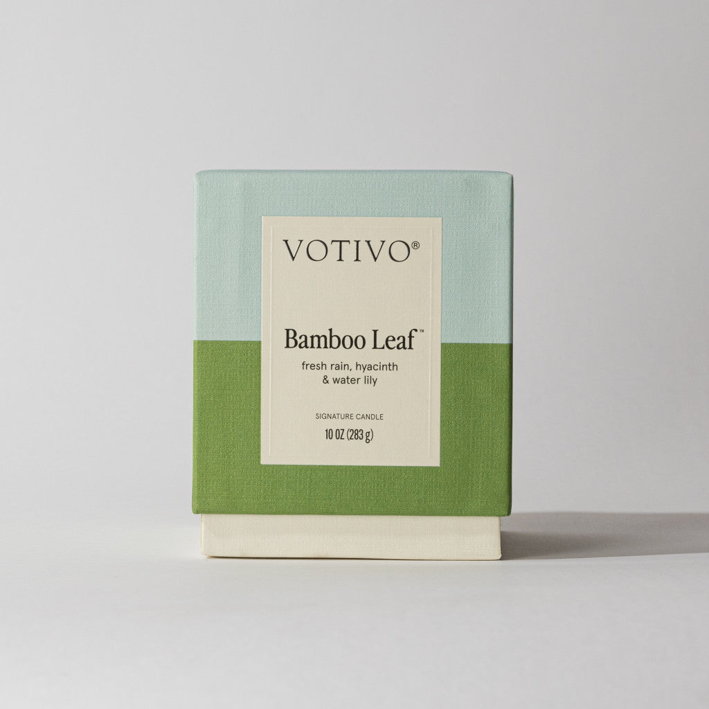 The Votivo Bamboo Leaf 10 oz. Signature Candle features a two-tone light blue and green box, crafted with soy blend wax, and offers notes of fresh rain, hyacinth, and water lily.