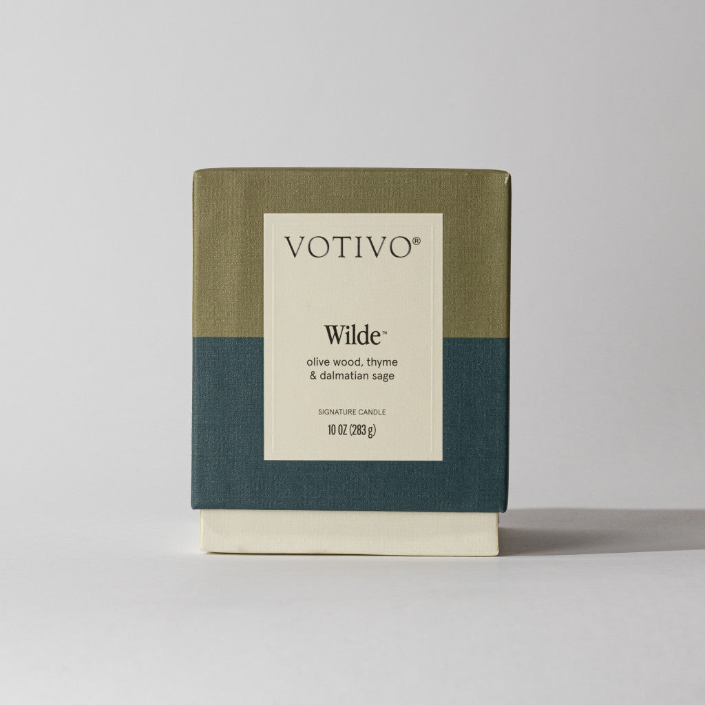 The Votivo Wilde 10 oz. Signature Candle features a soy blend wax and a unique olive wood, thyme, and dalmatian sage fragrance in an olive green, dark teal, and cream color block box on a plain background.