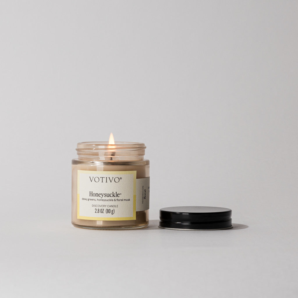 A Votivo Honeysuckle Discovery Candle burns in a glass jar on a white surface beside its black lid, filling the air with a soft floral scent.