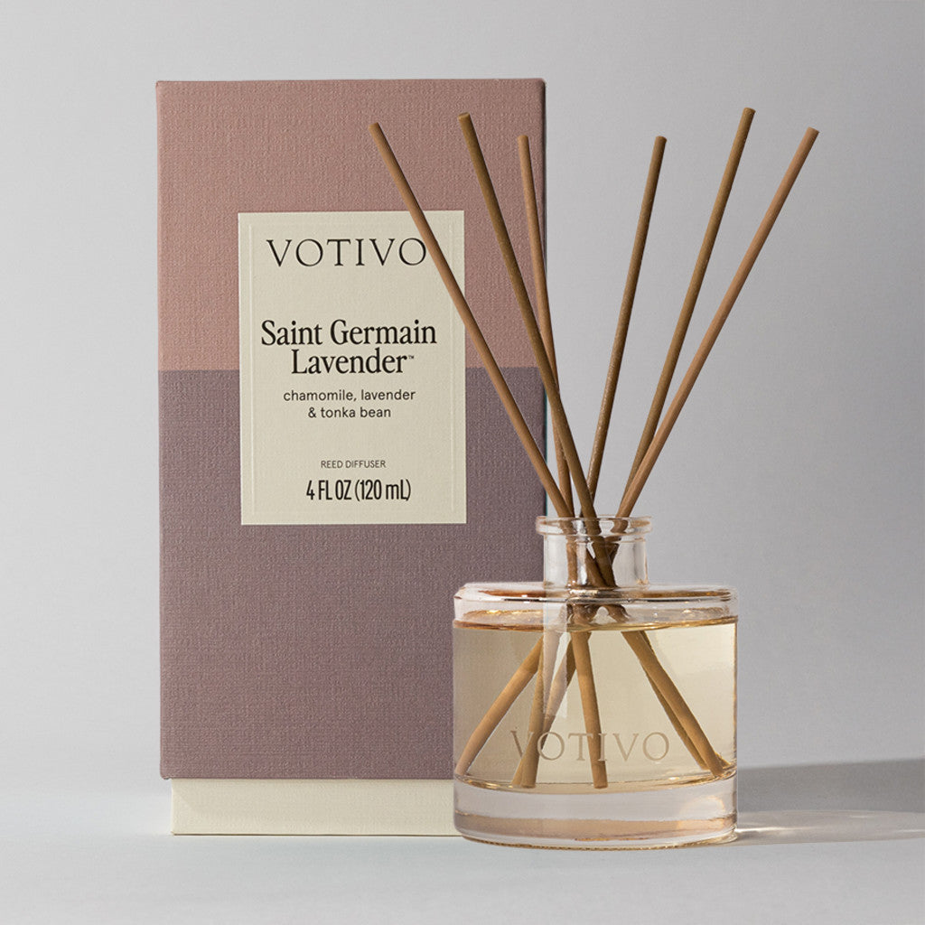 A Votivo Saint Germain Lavender Reed Diffuser with brown reeds in a clear glass bottle sits before its branded packaging featuring product details.