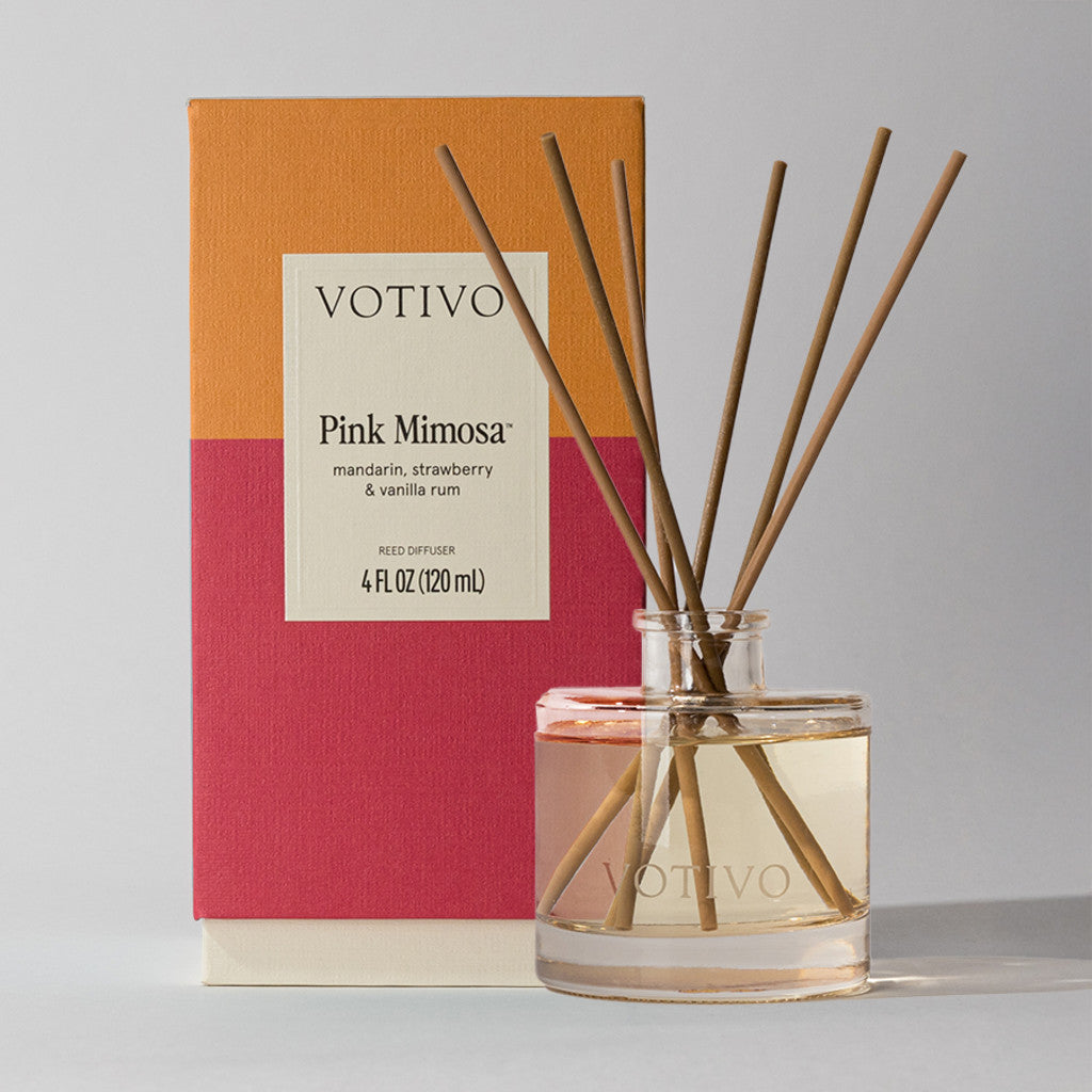 The Votivo Pink Mimosa Reed Diffuser, displayed with wooden sticks and its vibrant orange, red, and tan box, features a fruity fragrance blend of mandarin, strawberry, and vanilla rum.