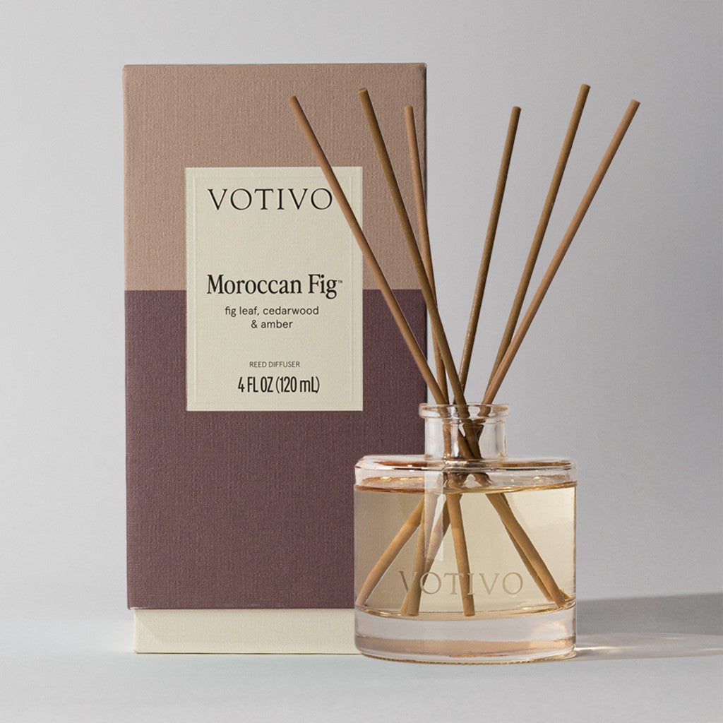 The Votivo Moroccan Fig Reed Diffuser fills your space with continuous fig leaf, cedarwood, and amber fragrance, featuring a clear glass bottle and reed sticks alongside its elegant beige and mauve packaging.