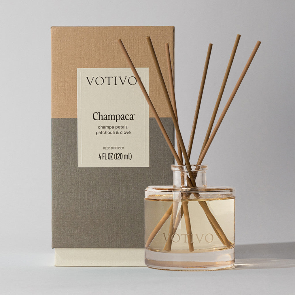 The Votivo Champaca Reed Diffuser features fiber reeds in a clear glass bottle, with a woody scent of champaca, patchouli & clove. Shown before its beige and gray box. 4 FL OZ (120 mL).