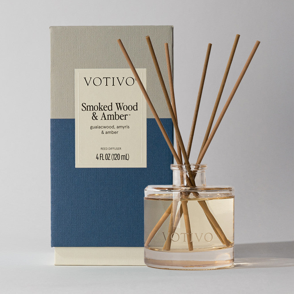 A Votivo Smoked Wood & Amber Reed Diffuser is placed before its box. The glass jar holds liquid and several reeds, while the box displays the Votivo brand, product name, and scent information.