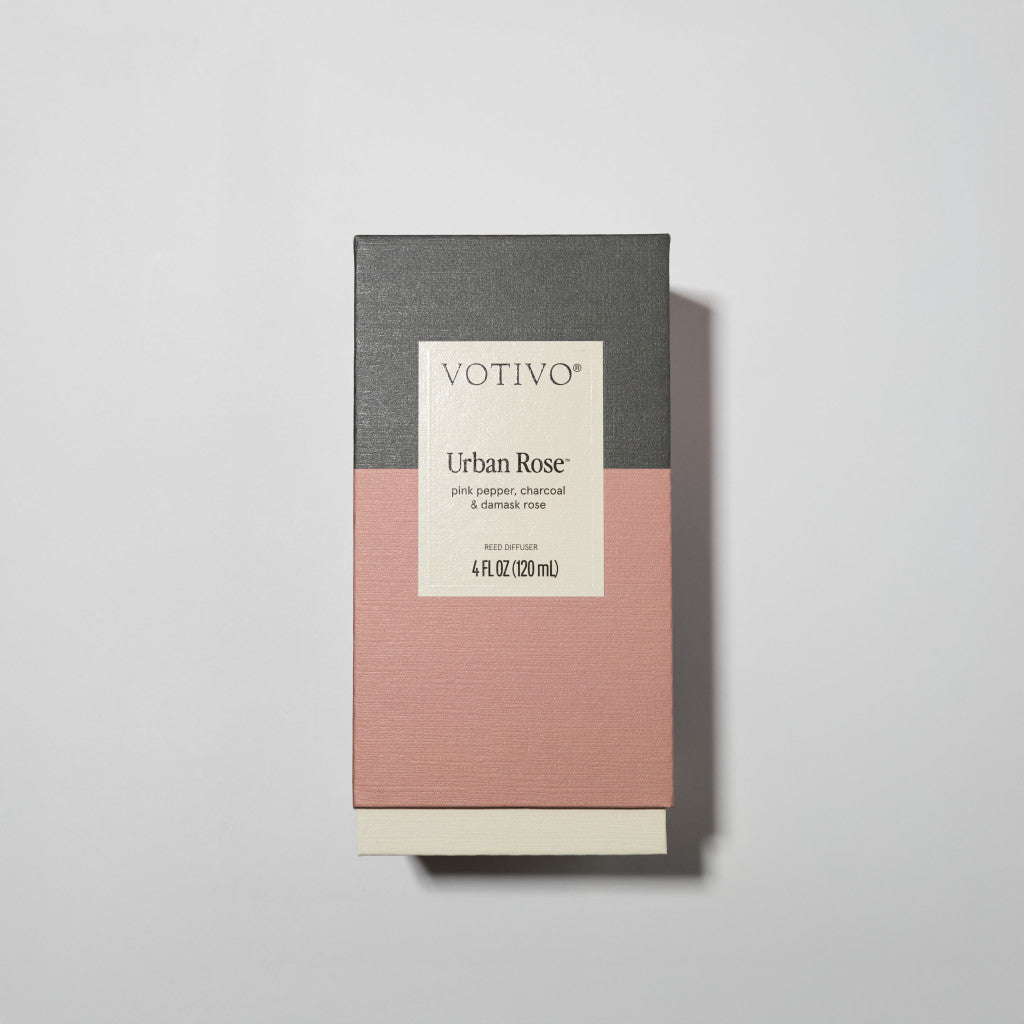 The Votivo Urban Rose Reed Diffuser features a gray and pink rectangular box and fiber reeds. Notes of pink pepper, charcoal, and Damask rose fill the 4 fl oz (120 mL) bottle for a modern floral experience.