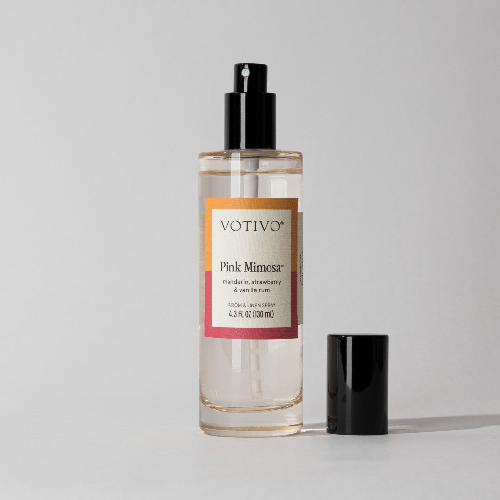 A clear Votivo Pink Mimosa Room & Linen Spray with a gold and pink label and black cap rests on a light surface. Its fruity scent instantly brightens any room—just one spritz is all it takes.