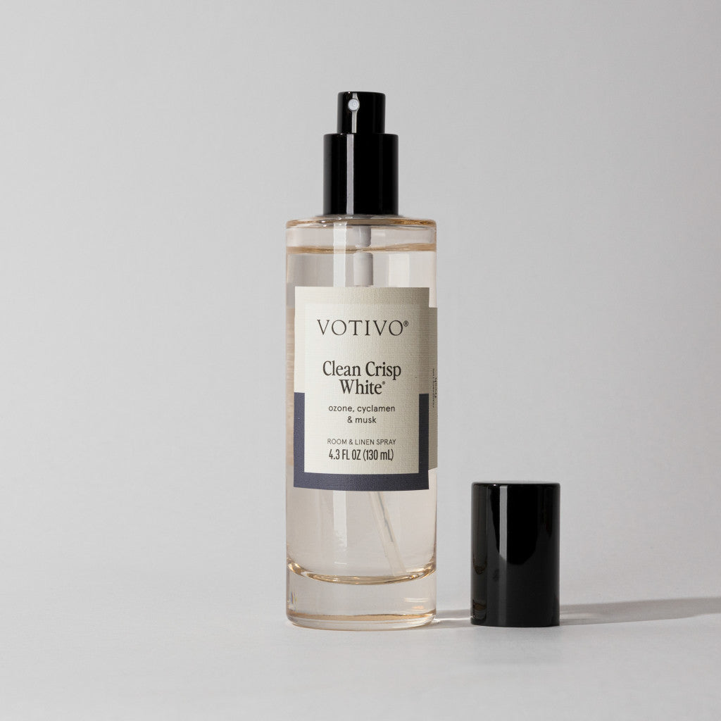 A bottle of Votivo Clean Crisp White Room & Linen Spray, featuring a black cap, sits on a simple light background. The label emphasizes this fresh scent, ideal for brightening any space.