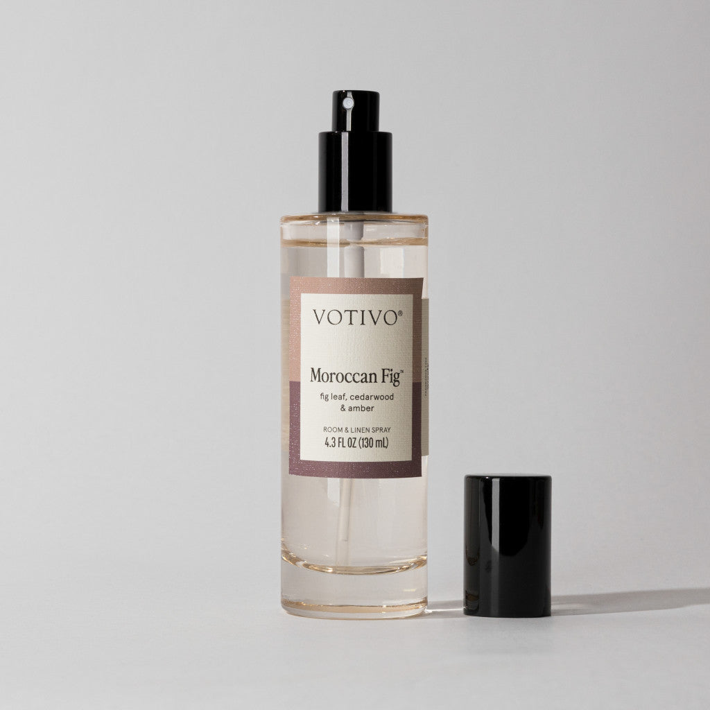 A clear spray bottle labeled Votivo Moroccan Fig Room & Linen Spray, featuring a black nozzle and a matching cap placed beside it, stands against a simple, light background.