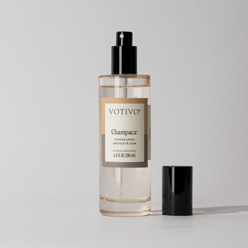 A clear bottle of Votivo Champaca Room & Linen Spray with a black nozzle and separate cap is set against a light background, highlighting its elegant champaca scent with subtle patchouli notes.