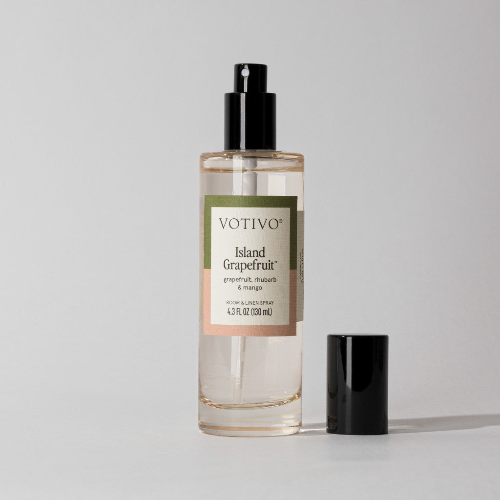 A clear bottle of Votivo Island Grapefruit Room & Linen Spray features a pink and green label, black spray nozzle and cap, all set against a plain light background. Experience the refreshing citrus scent with every spritz.