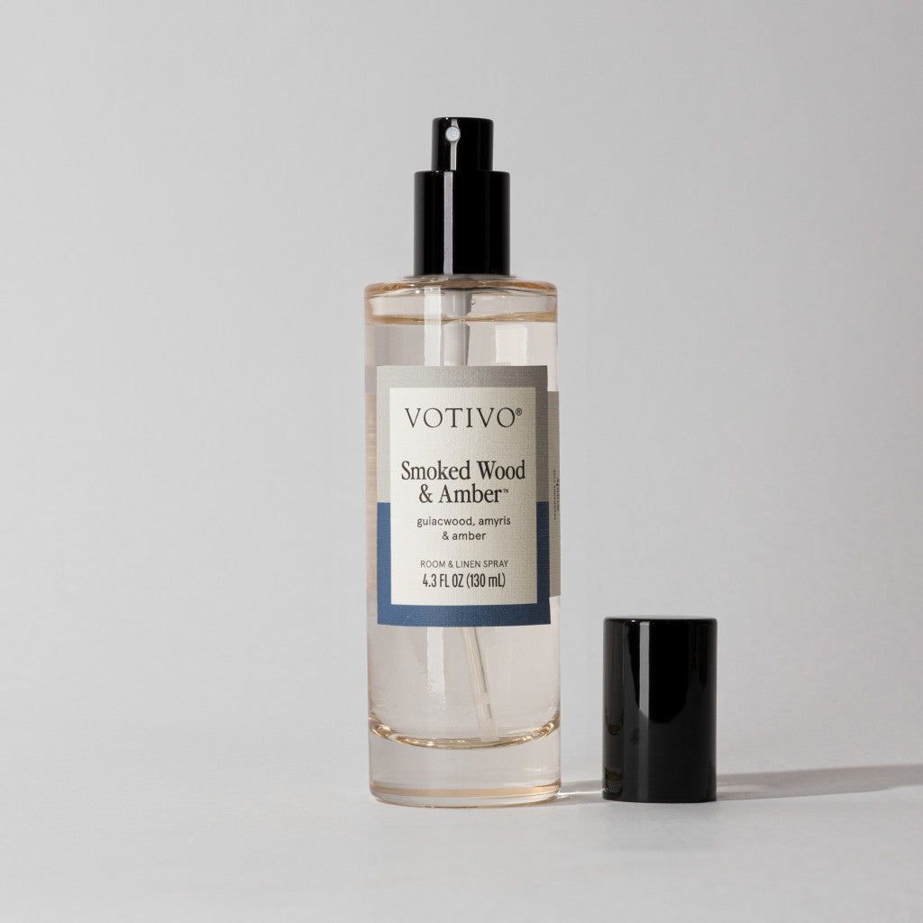 A clear spray bottle of Votivo Smoked Wood & Amber Room & Linen Spray, featuring a black nozzle and cap, sits on a surface against a light gray background.