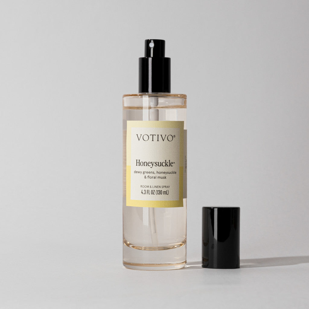 A clear Votivo Honeysuckle Room & Linen Spray bottle with a black nozzle and cap rests on a light gray background, evoking the fresh floral fragrance of honeysuckle.