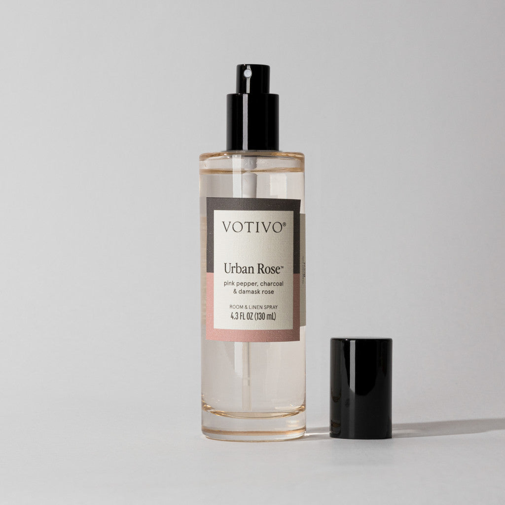 A clear bottle of Votivo Urban Rose Room & Linen Spray with a black nozzle and cap sits on a light gray background. The label notes charcoal rose, pink pepper, and damask rose in this distinctive urban fragrance.